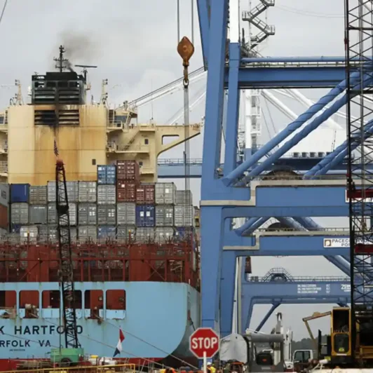 Cargo ship loaded with shipping containers docked beneath large industrial port cranes.