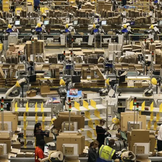 Wide view of a busy fulfillment center with workers, conveyor stations, and stacks of cardboard boxes.