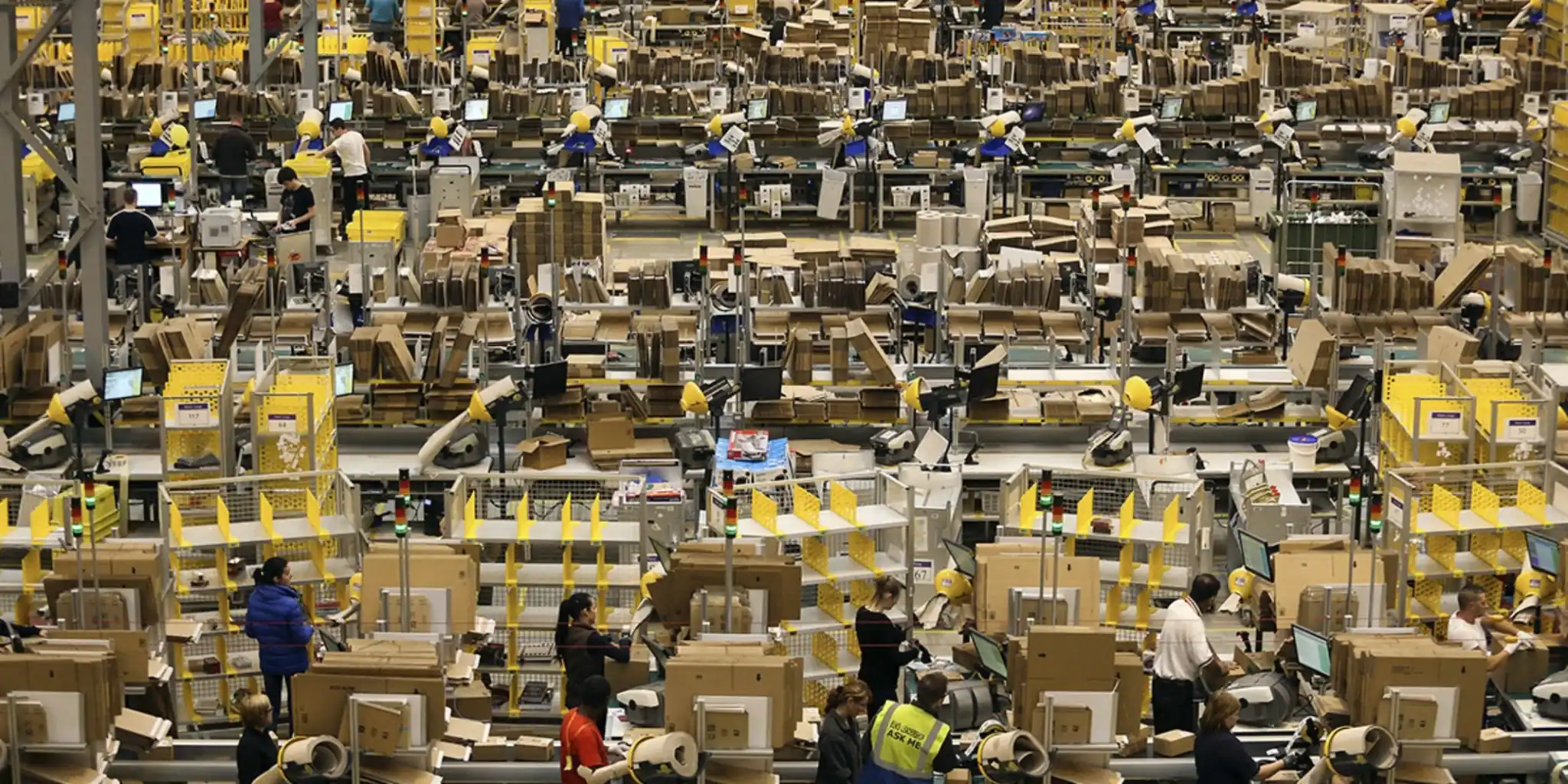 Wide view of a busy fulfillment center with workers, conveyor stations, and stacks of cardboard boxes.