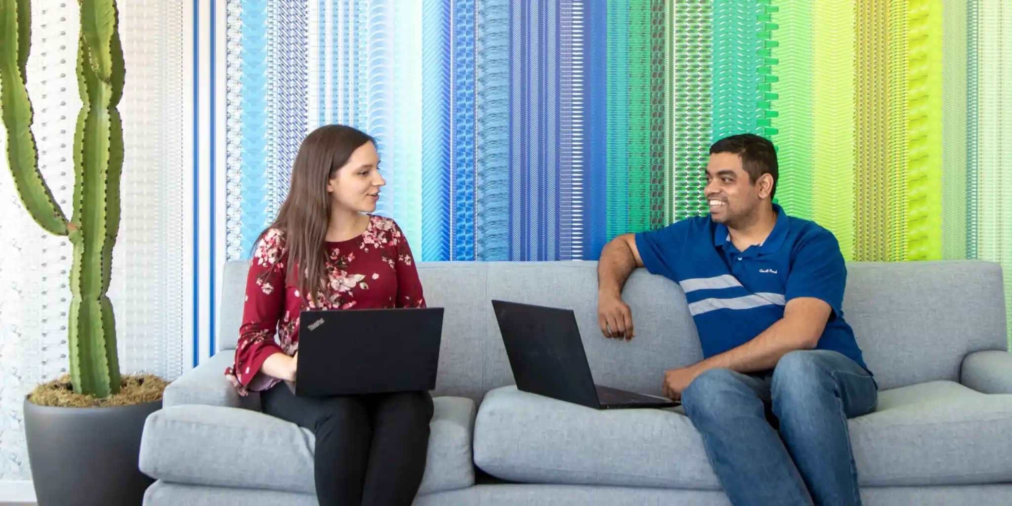 Two coworkers with laptops talking on a couch in front of a colorful textured wall.