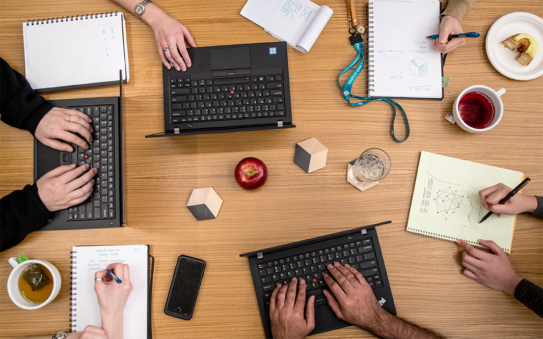 Picture of a tabletop with three computers and three notebooks with people working