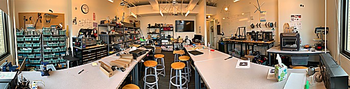 Panoramic view of a maker workshop lab with workbenches, tool storage bins, 3D printers, and stools.