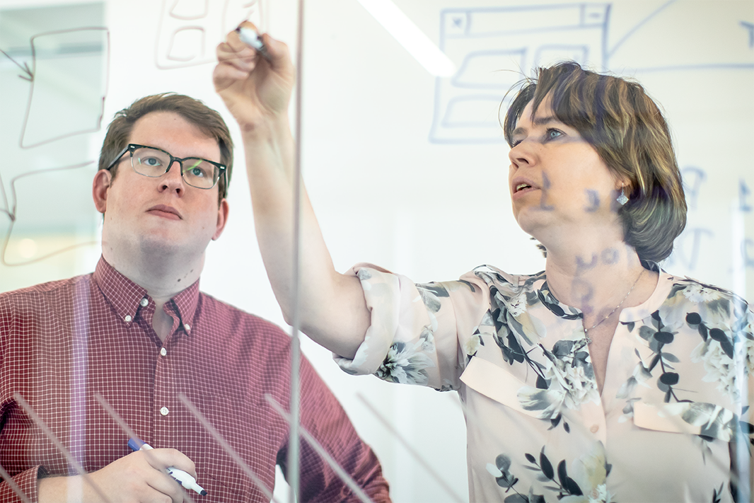 Man and a woman working out a complex problem together on a glass dry erase board