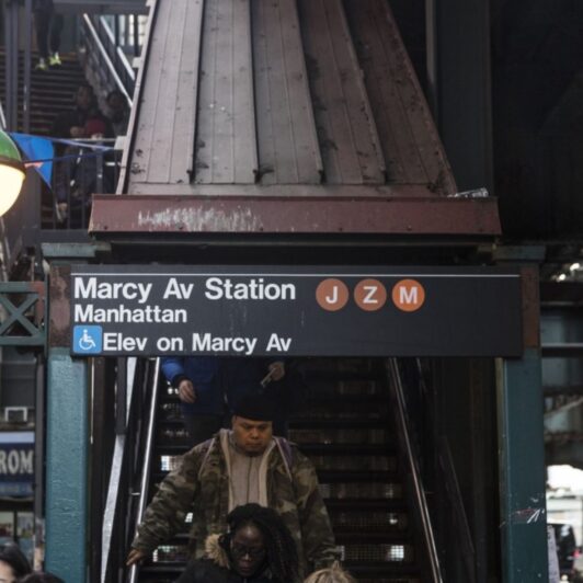 Outdoor stairwell leading to the NYC subway with a sign displaying that there is also an elevator available on the avenue.