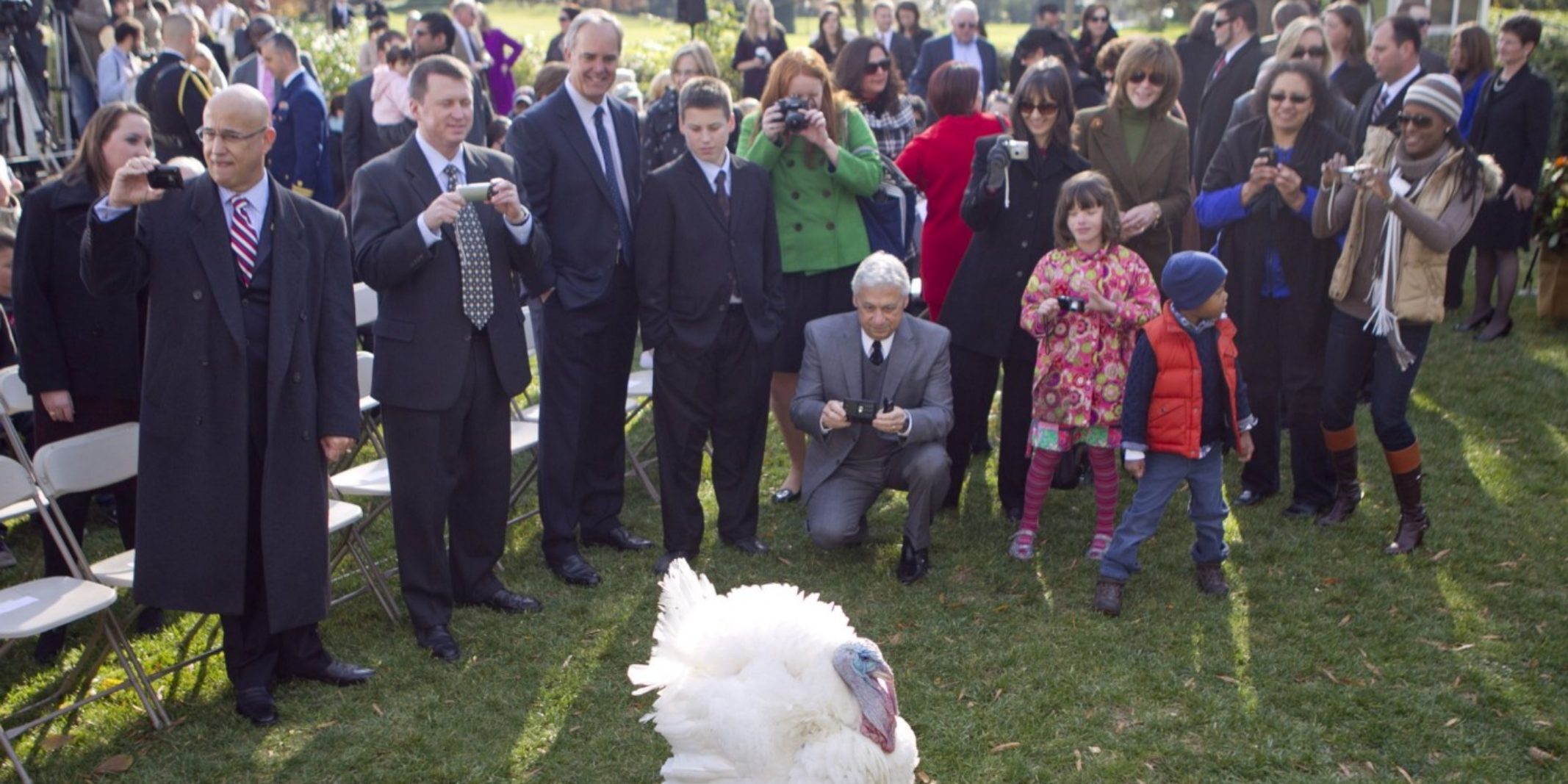 A crowd of smiling families gather around a live turkey, taking photos.