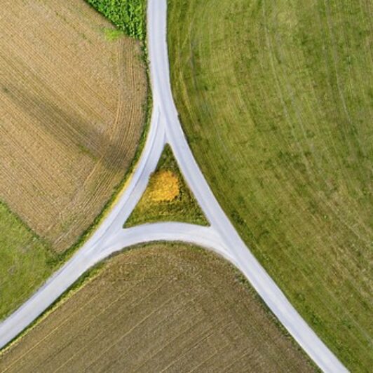 Aerial view of farmland fields in green and brown patches converging around a Y-shaped road intersection.