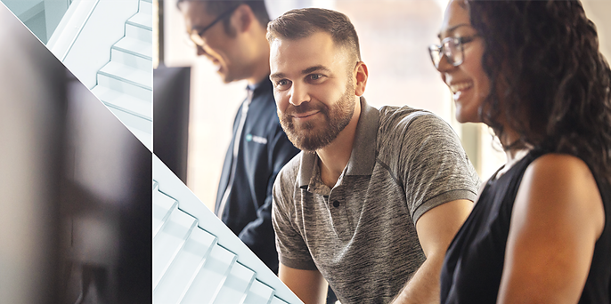 A bearded man in a gray polo smiles while talking with two colleagues in a bright office setting.