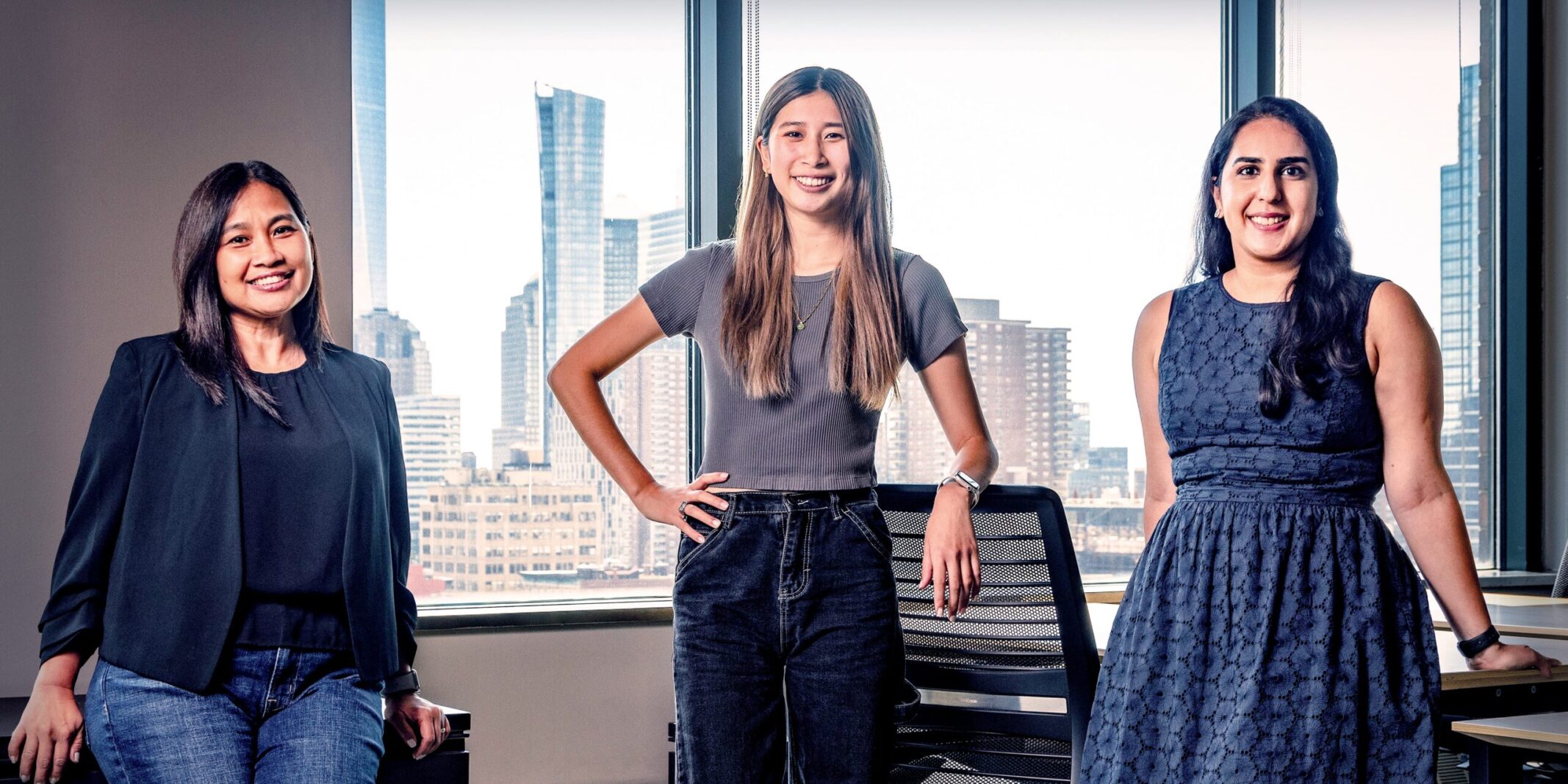 In an office, three woman stand confidently: Mae, Head of Critical Applications and Data Reliability Engineering, Katherine, a Software Engineer, and Aditi, an Application Security Engineer.