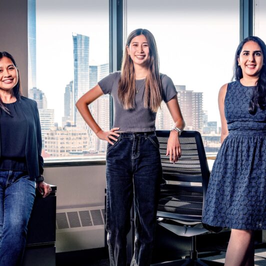 In an office, three woman stand confidently: Mae, Head of Critical Applications and Data Reliability Engineering, Katherine, a Software Engineer, and Aditi, an Application Security Engineer.