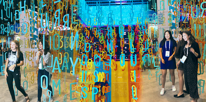 Conference attendees walk past a colorful hanging letter installation and a lit sign at an event booth.
