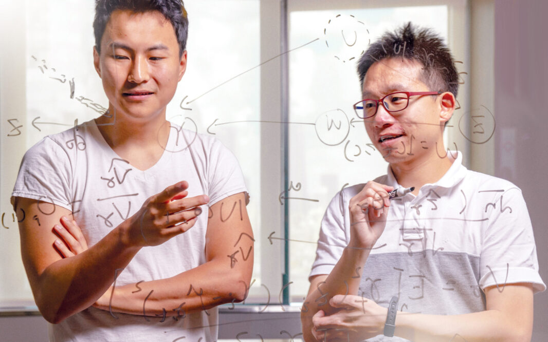 Two graduate students write equations on a glass whiteboard.