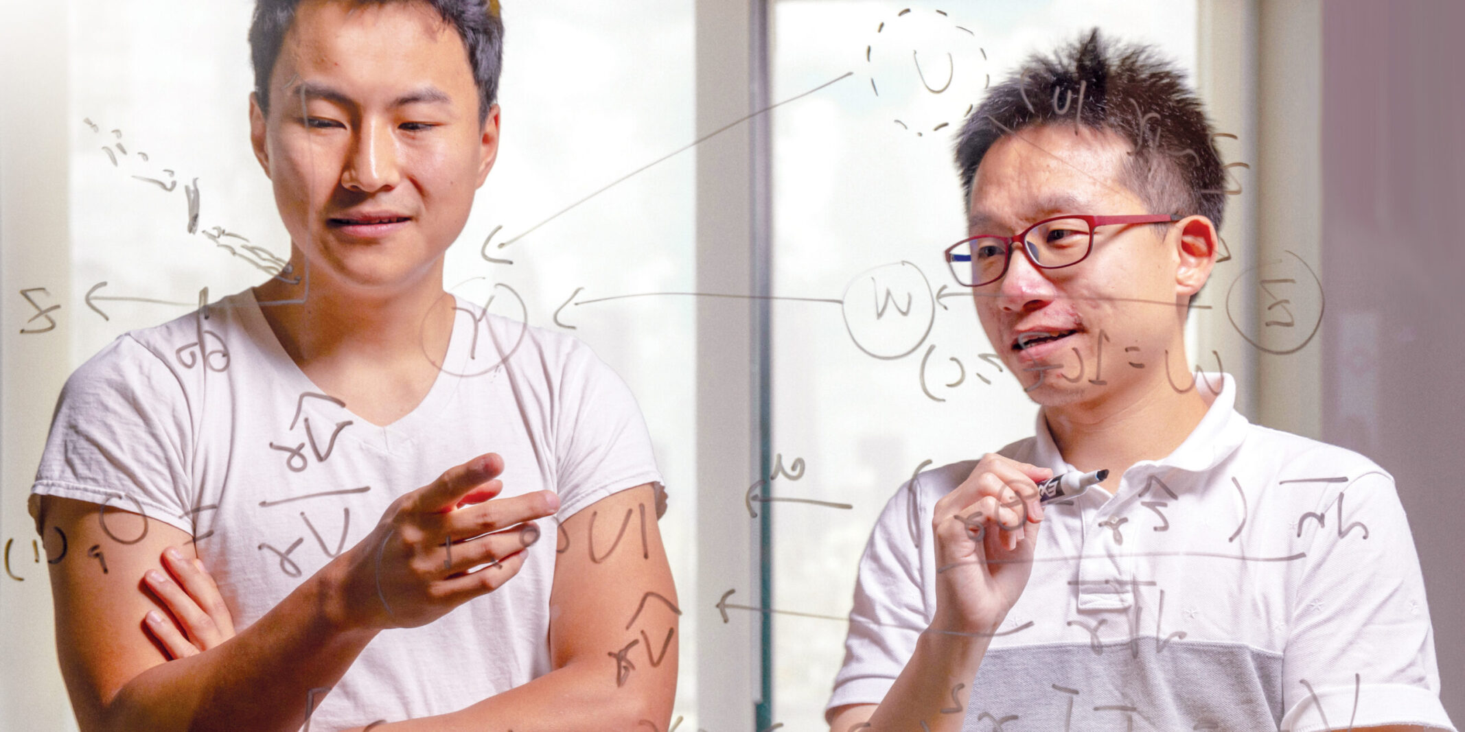 Two graduate students write equations on a glass whiteboard.