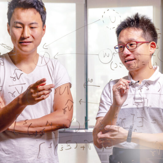 Two graduate students write equations on a glass whiteboard.