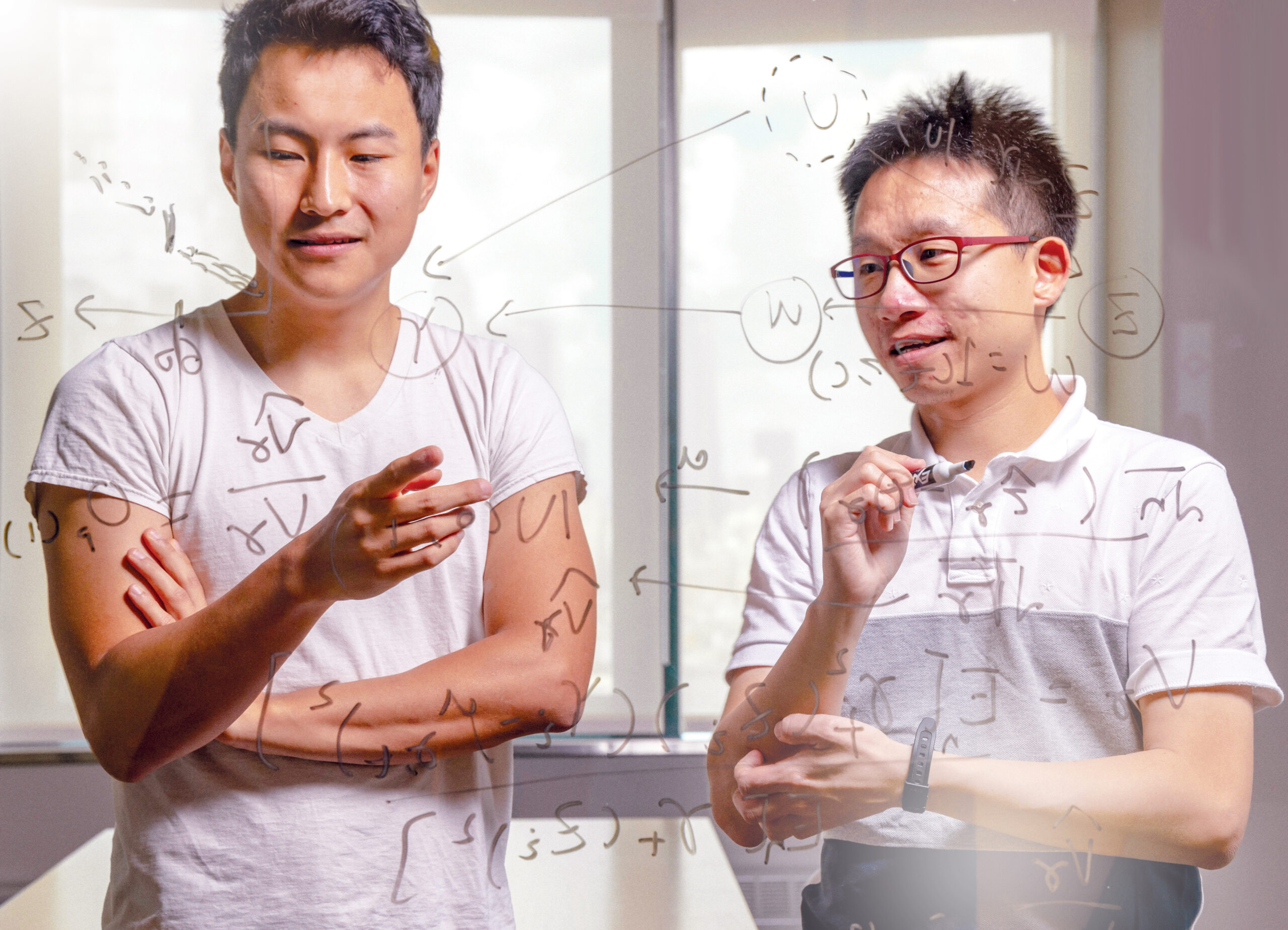 Two graduate students write equations on a glass whiteboard.