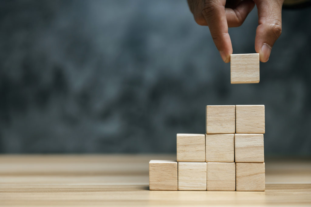 Hand putting blank wooden cube block on a staircase-like stack of blocks on dark background