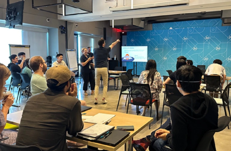 Group workshop in a classroom with participants seated at tables while presenters speak at the front.