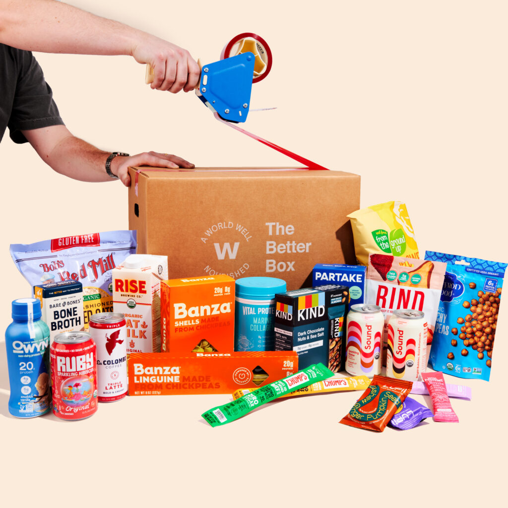 A brown cardboard box featuring the Wellfare Better Box logo is being taped shut, with examples of packaged food items from the box in front of it.