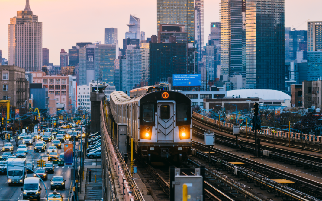 A subway train moves toward the photographer on an elevated subway line with a background of an urban landscape of highrises, and a road full of car traffic below and parallel to the subway line on the left.