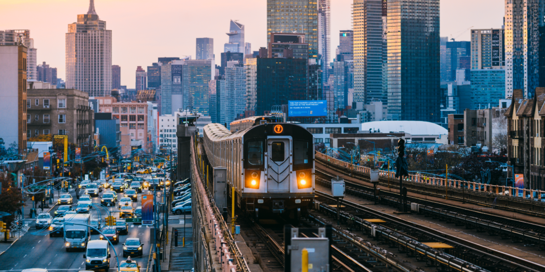 A subway train moves toward the photographer on an elevated subway line with a background of an urban landscape of highrises, and a road full of car traffic below and parallel to the subway line on the left.