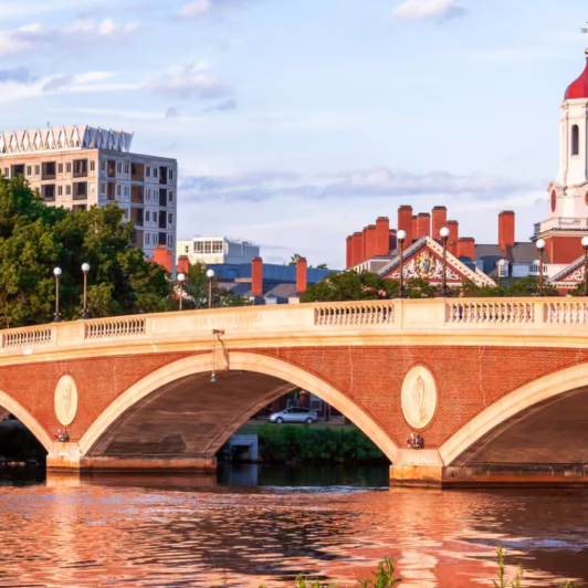 Harvard campus buildings and a red-brick arched bridge spanning the Charles River under a partly cloudy sky.