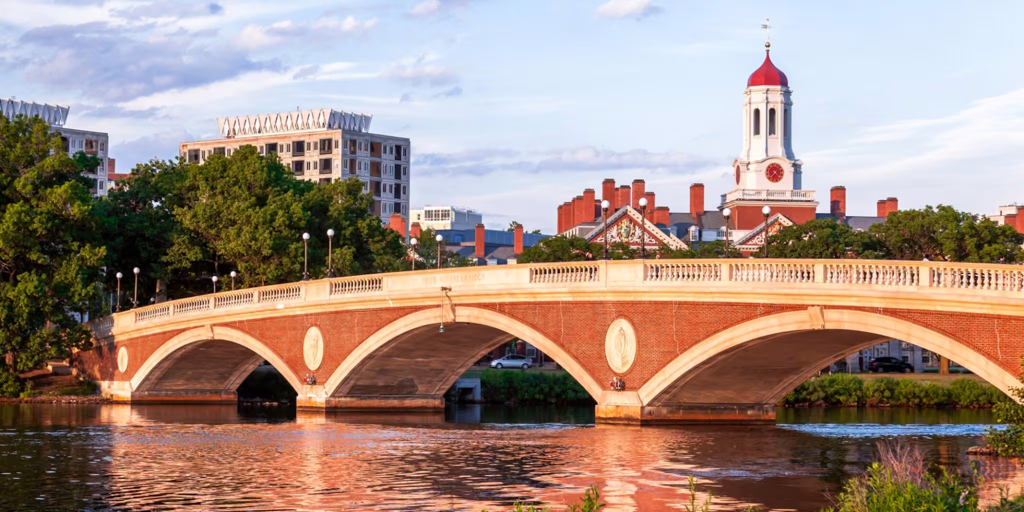 Harvard campus buildings and a red-brick arched bridge spanning the Charles River under a partly cloudy sky.