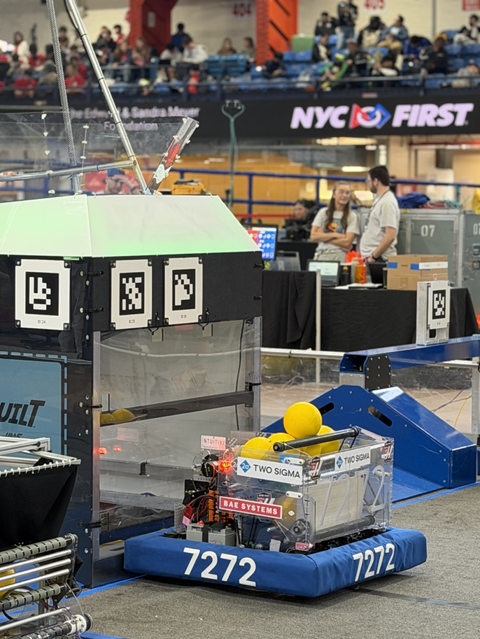 A competition robot with QR-style markers and yellow balls sits at an NYC FIRST robotics event, sponsor logos and crowd visible behind it.