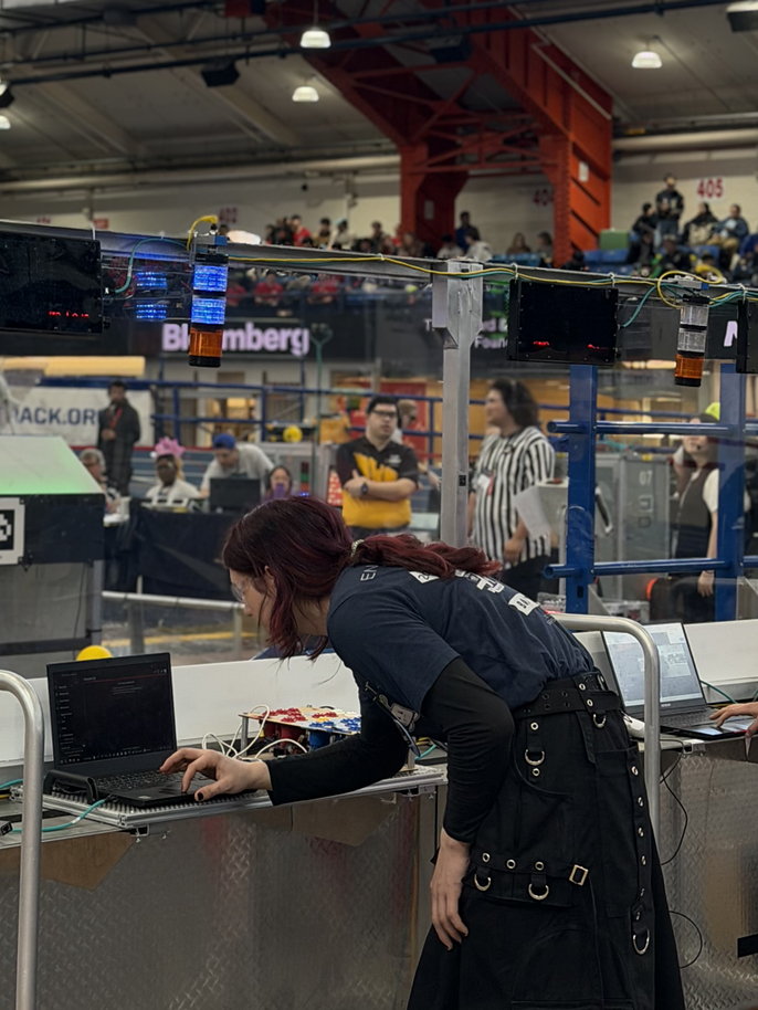 A woman with red hair leans over a laptop at a robotics competition arena, spectators and a Bloomberg sign visible behind her.
