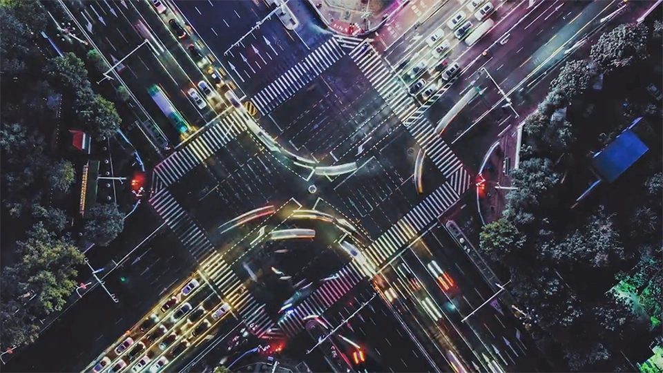 Aerial nighttime view of a large city intersection with moving traffic, crosswalks, and surrounding trees.