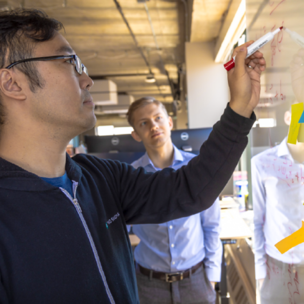 Man writing equations on a glass whiteboard while two colleagues look on in an office setting.
