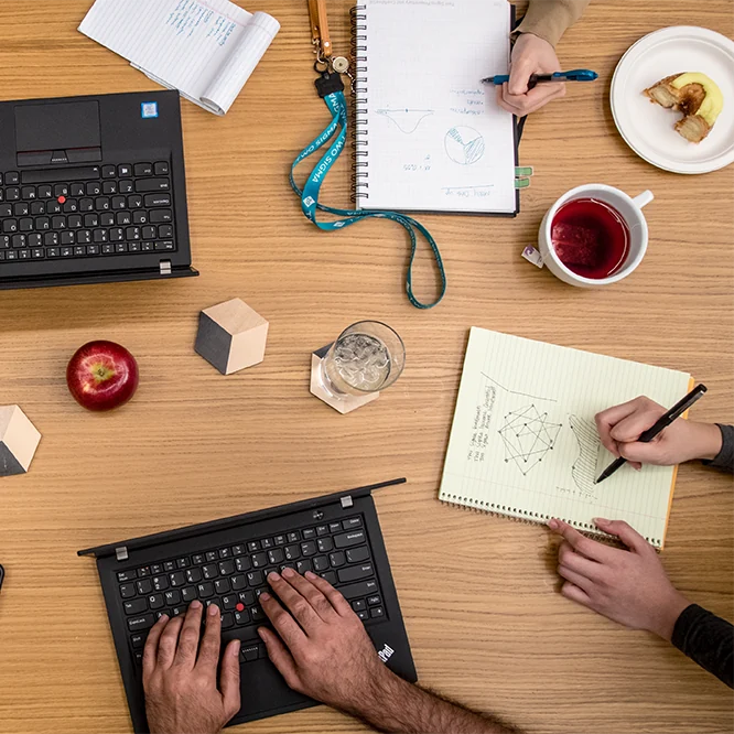 Top-down view of a shared worktable with laptops, notebooks, charts, drinks, and hands working together.