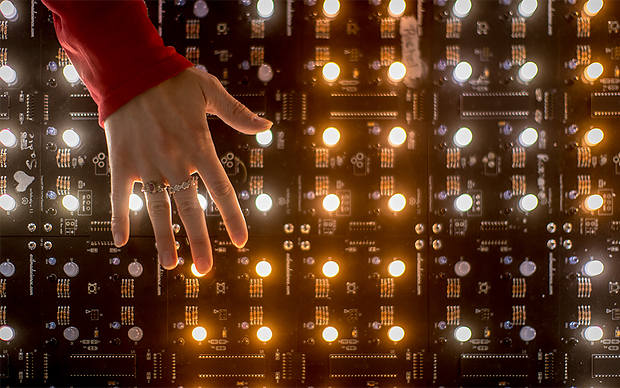 A hand with rings reaches toward a glowing circuit board covered in lit components.