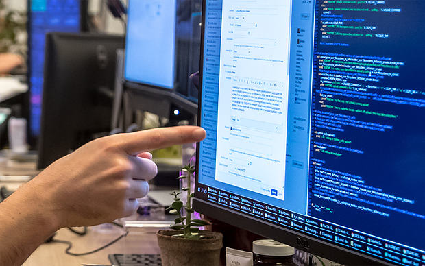 A hand points at a computer monitor displaying a form and lines of code, with a small plant nearby.