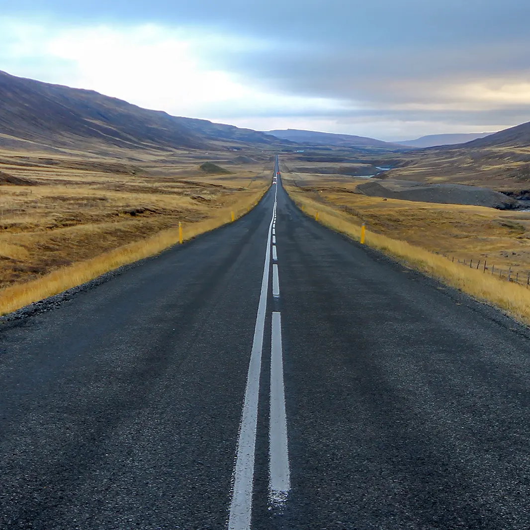 A long, empty two-lane road stretches through a golden-brown valley between mountains under a cloudy sky.