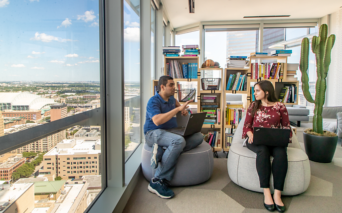 A man and woman sit in lounge chairs by floor-to-ceiling windows overlooking a city skyline, each working on a laptop beside a tall bookshelf.