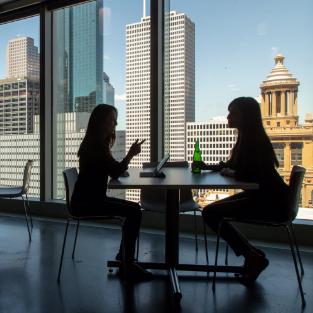 Two women in silhouette sit across a small table by a window overlooking the Houston skyline and a domed clock tower building.