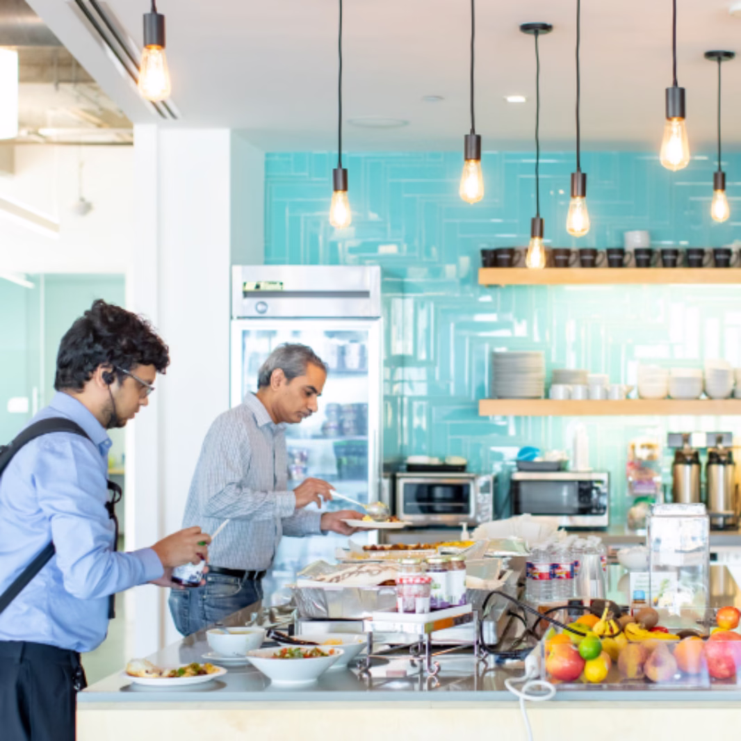 Two people serving themselves food at a buffet counter in a modern office kitchen with hanging lights and turquoise walls.