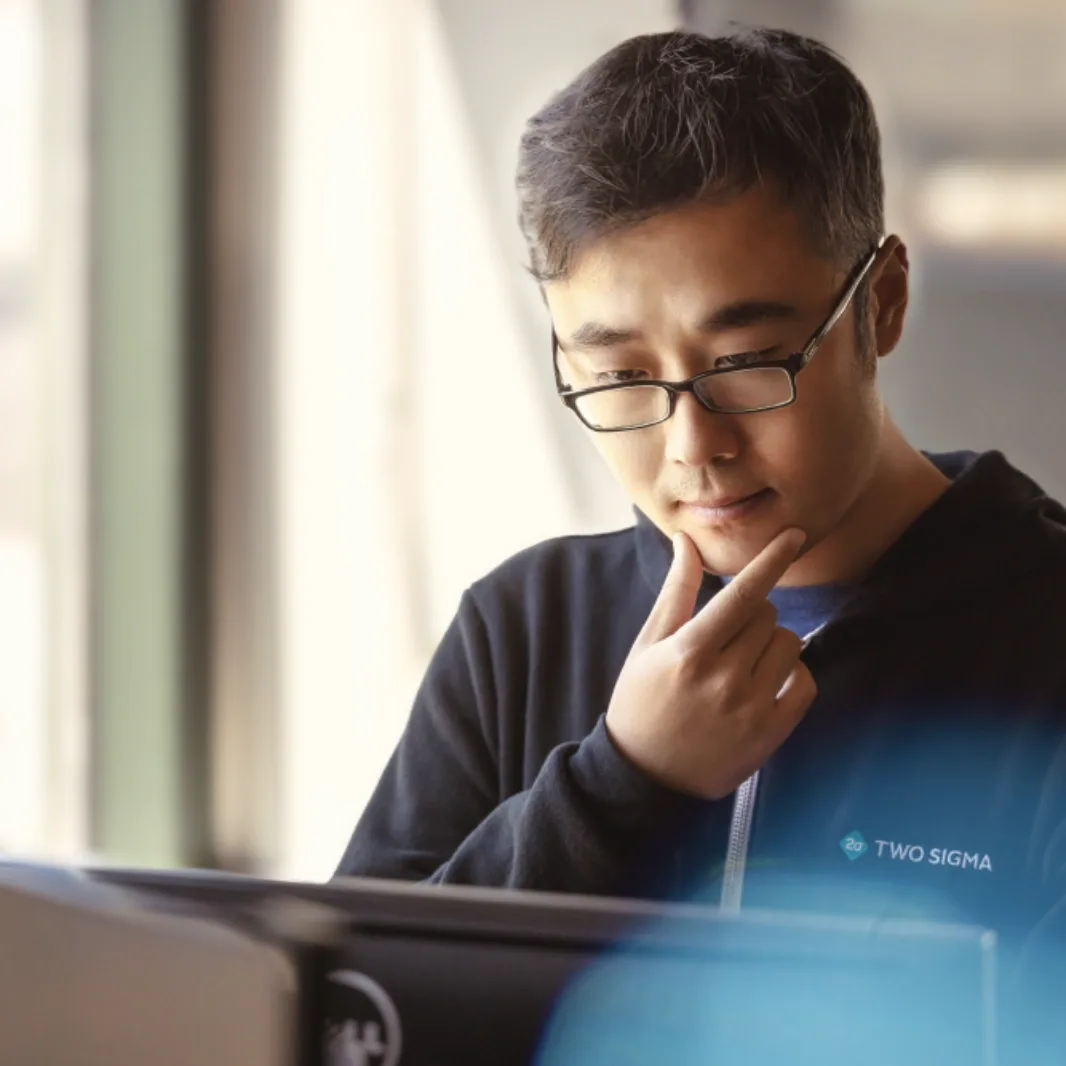 Person wearing glasses and a Two Sigma hoodie looking thoughtfully at a laptop.