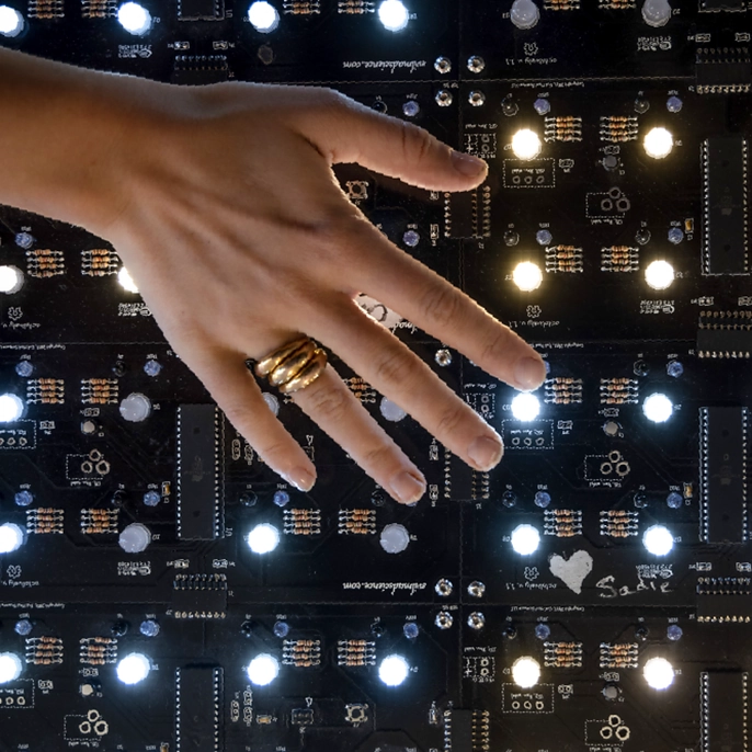 A hand wearing rings reaches over an illuminated electronic circuit board panel dotted with lit indicator lights.