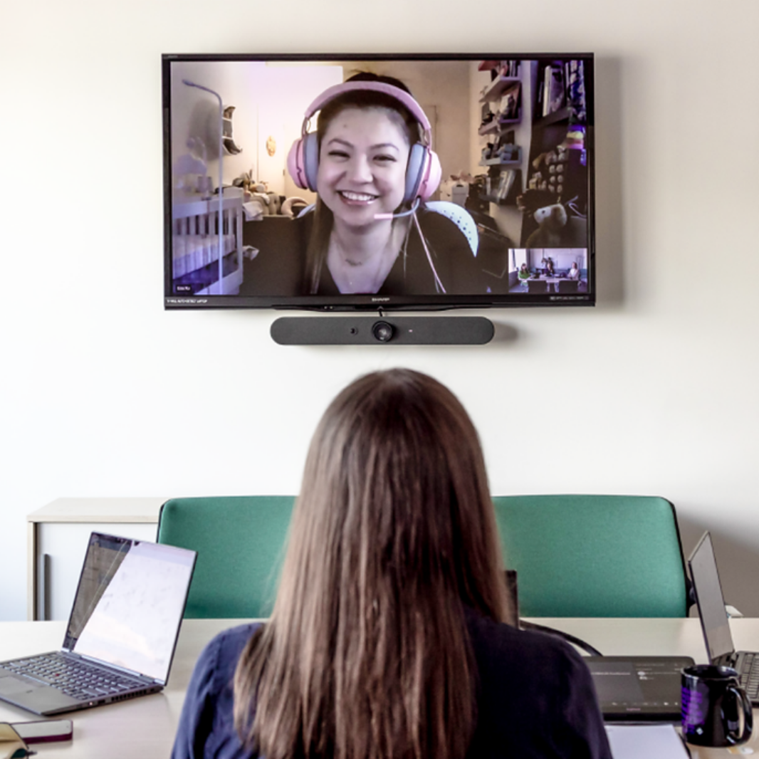 A woman views a wall-mounted screen showing a smiling colleague wearing headphones during a video call, seated at a laptop in a meeting room.