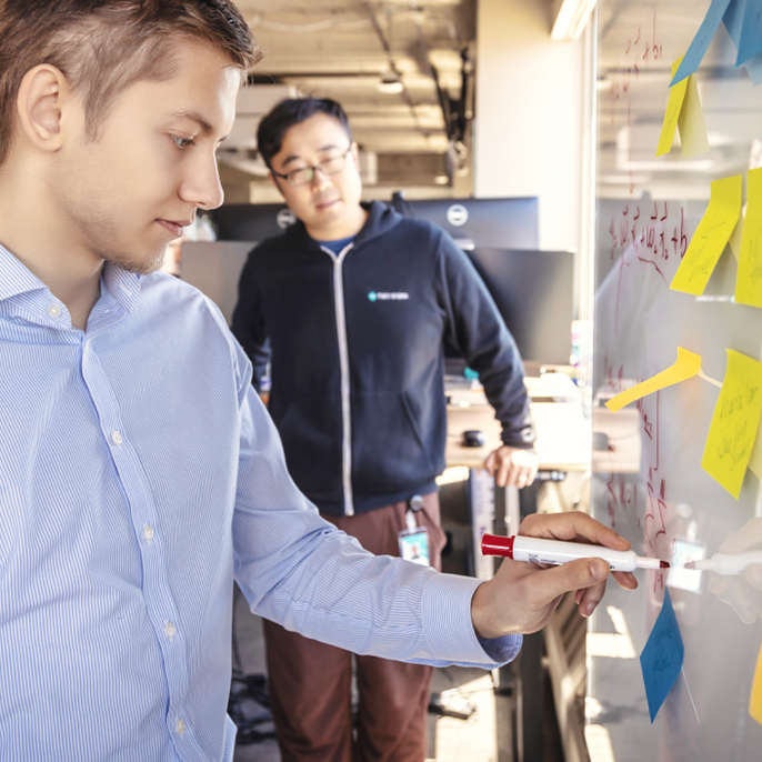 A man writes on a glass whiteboard covered in colorful sticky notes and equations while a colleague looks on in an open office.