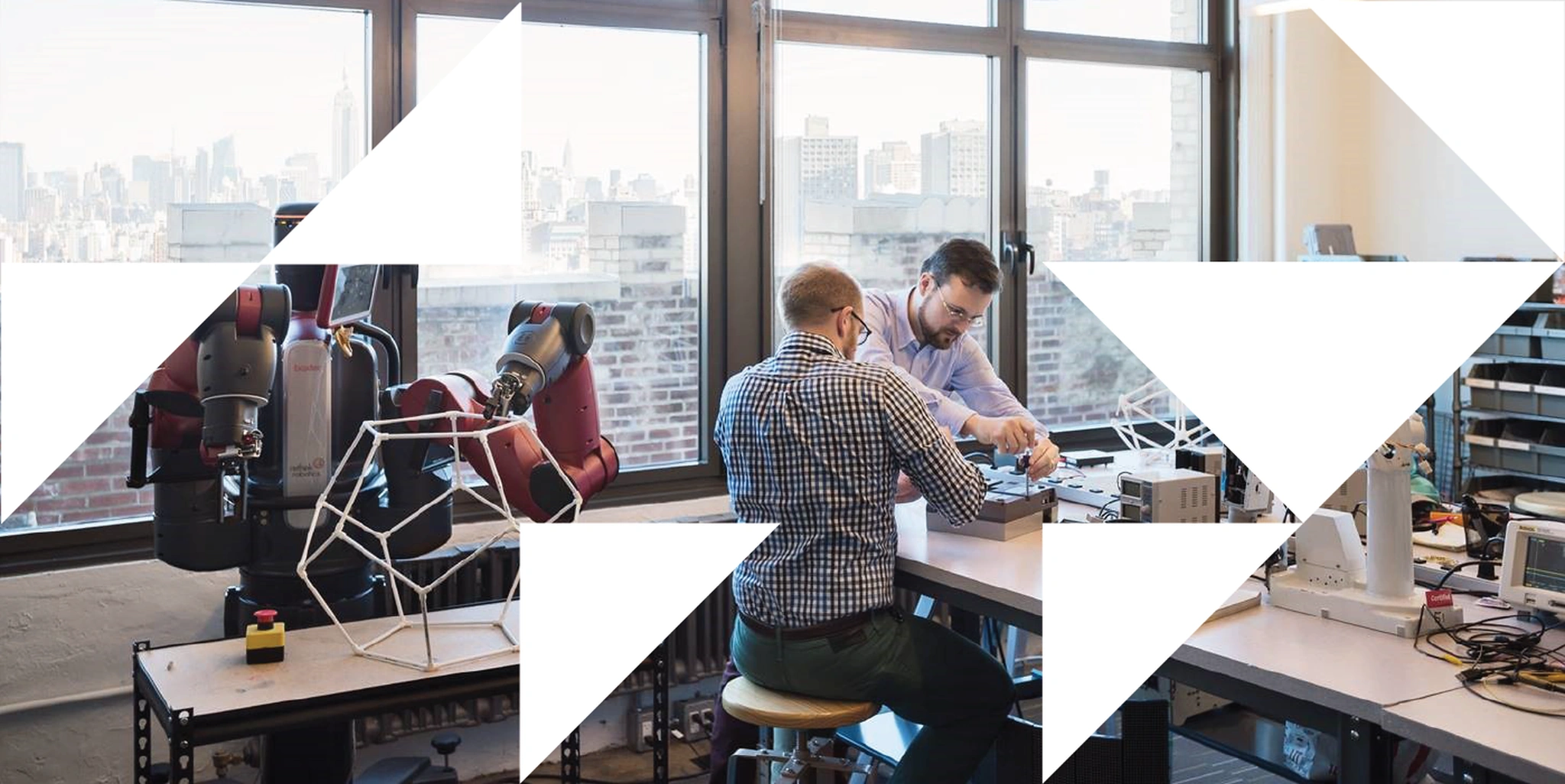 Two engineers work at a lab bench with a red robotic arm and 3D-printed geometric shapes, city skyline visible through windows, over a geometric triangle overlay.