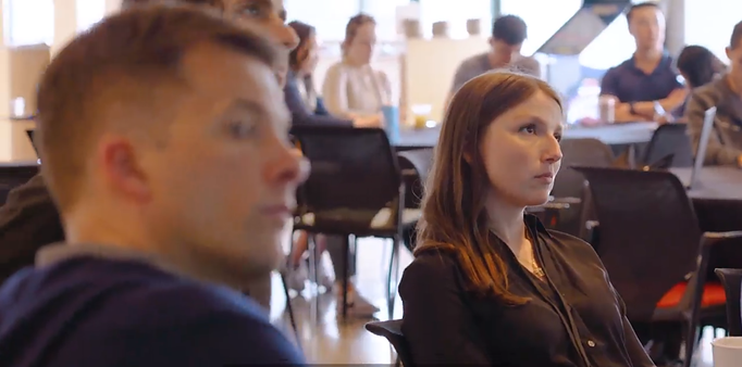 People seated and listening intently in a bright office space, with colleagues working at tables in the blurred background.